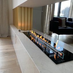 Dom&Greg - Nowoczesny biokominek z paleniskiem w obudowie z białego marmuru, z widocznym sterownikiem, na tle drewnianej podłogi i zasłon w salonie.