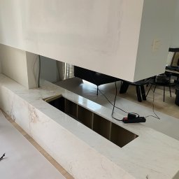 Dom&Greg - Nowoczesny kominek z marmurowym wykończeniem w trakcie montażu, zabezpieczony folią malarską, widoczne kable i listwa zasilająca, w tle salon.