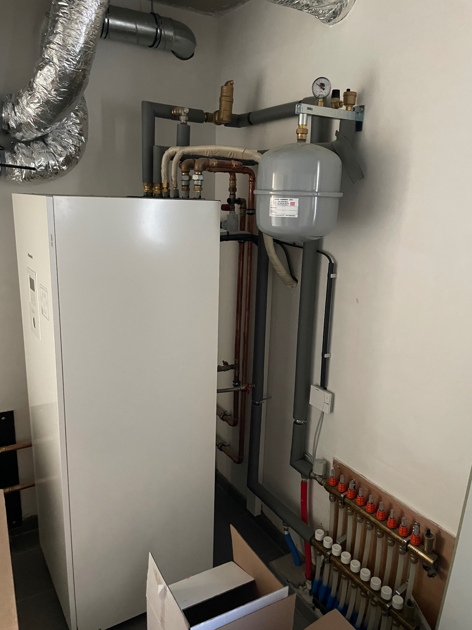 Pompa ciepła powietrze-woda z szarym zbiornikiem wyrównawczym, licznymi rurami miedzianymi i szarymi, oraz rozdzielaczem podłogowym z czerwonymi i białymi zaworami, zamontowana w pomieszczeniu...