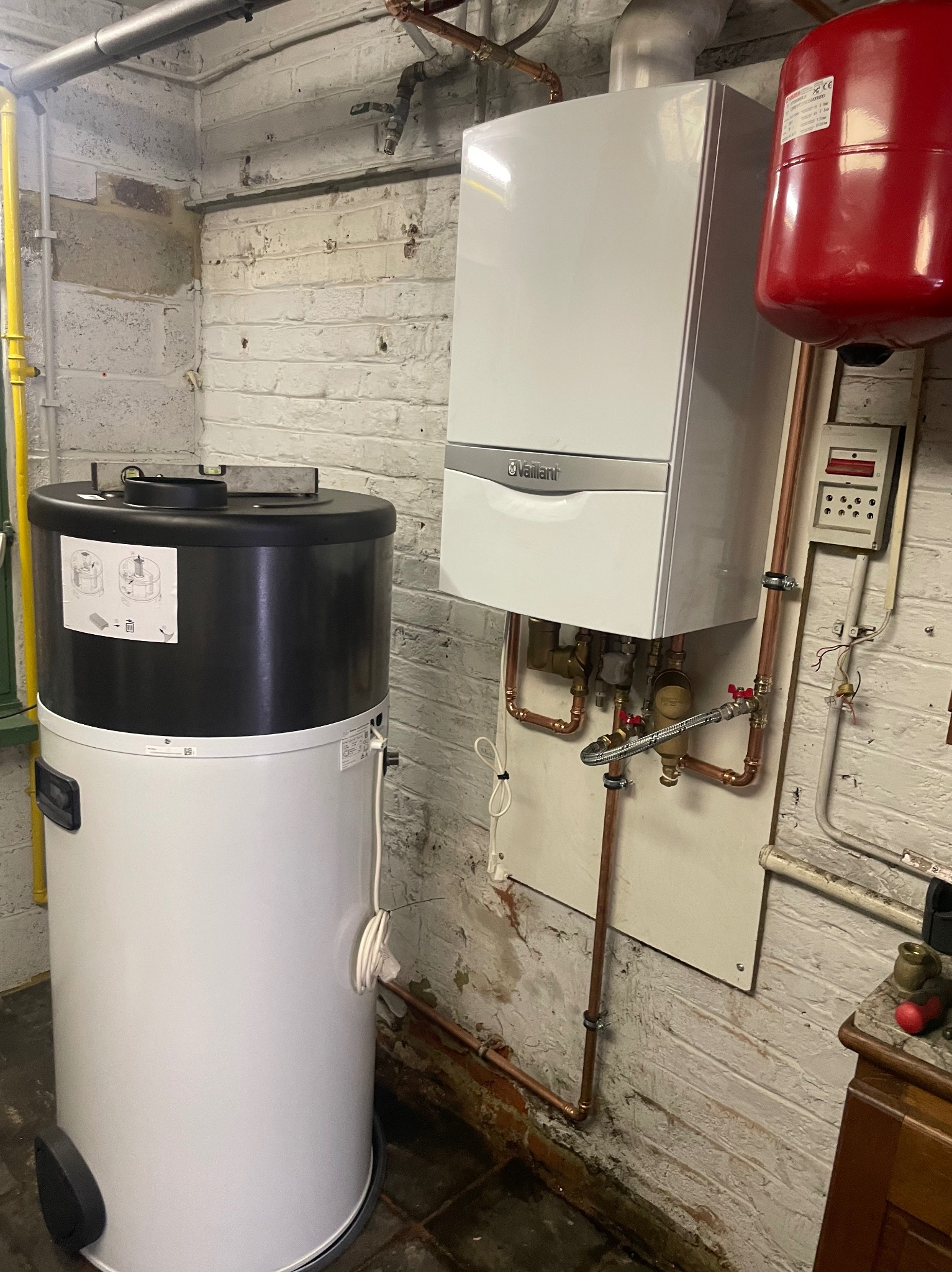 Nowoczesny, biały piec gazowy Vaillant zamontowany na pomalowanej na biało ceglanej ścianie, obok stoi biały zasobnik ciepłej wody z czarną górą, widoczne rury miedziane i instalacja elektryczna.
