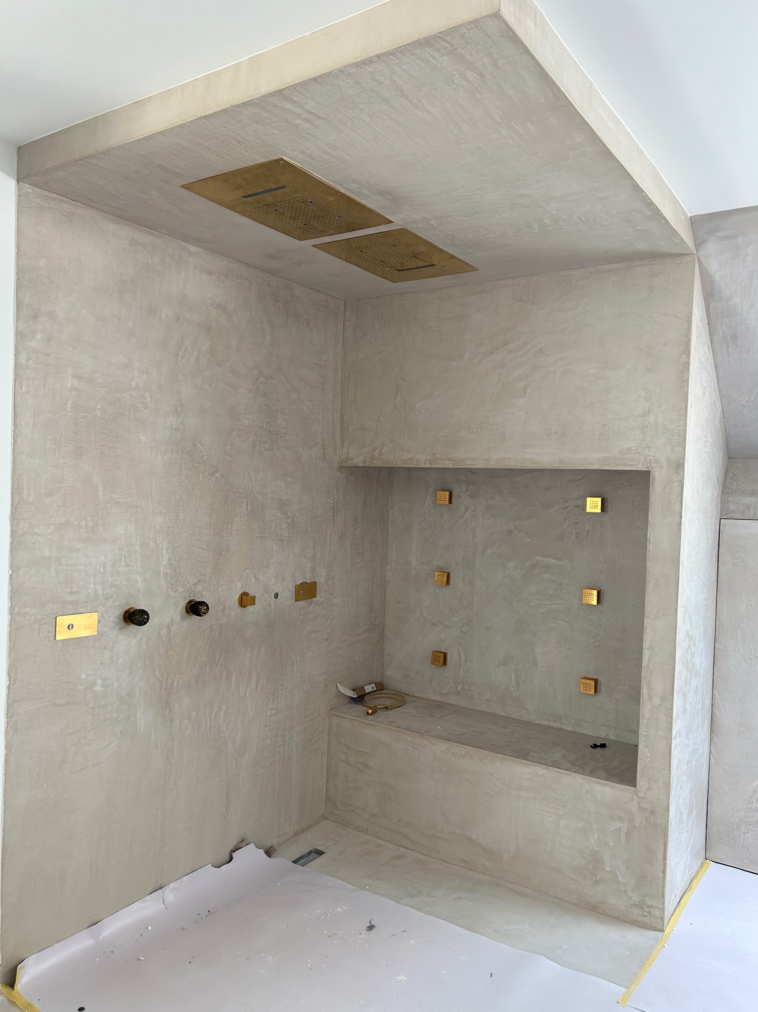 Łazienka w trakcie remontu: surowe ściany z efektem betonu, złote elementy armatury i deszczownicy, zabezpieczona podłoga.