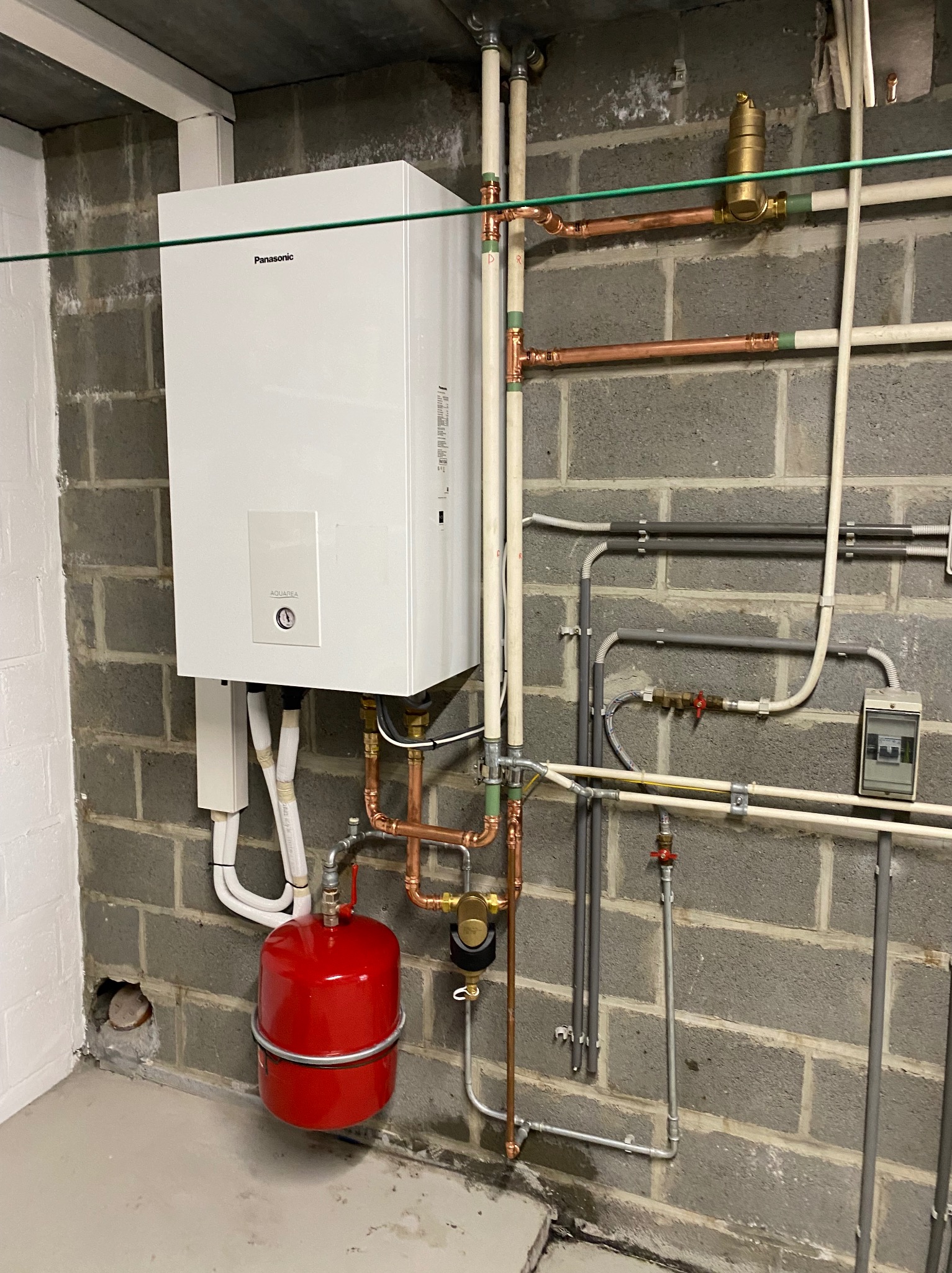 Biały piec gazowy marki Panasonic z czerwonym zbiornikiem wyrównawczym, zamontowany na ścianie z szarych betonowych bloczków, z widocznymi rurami miedzianymi i plastikowymi.