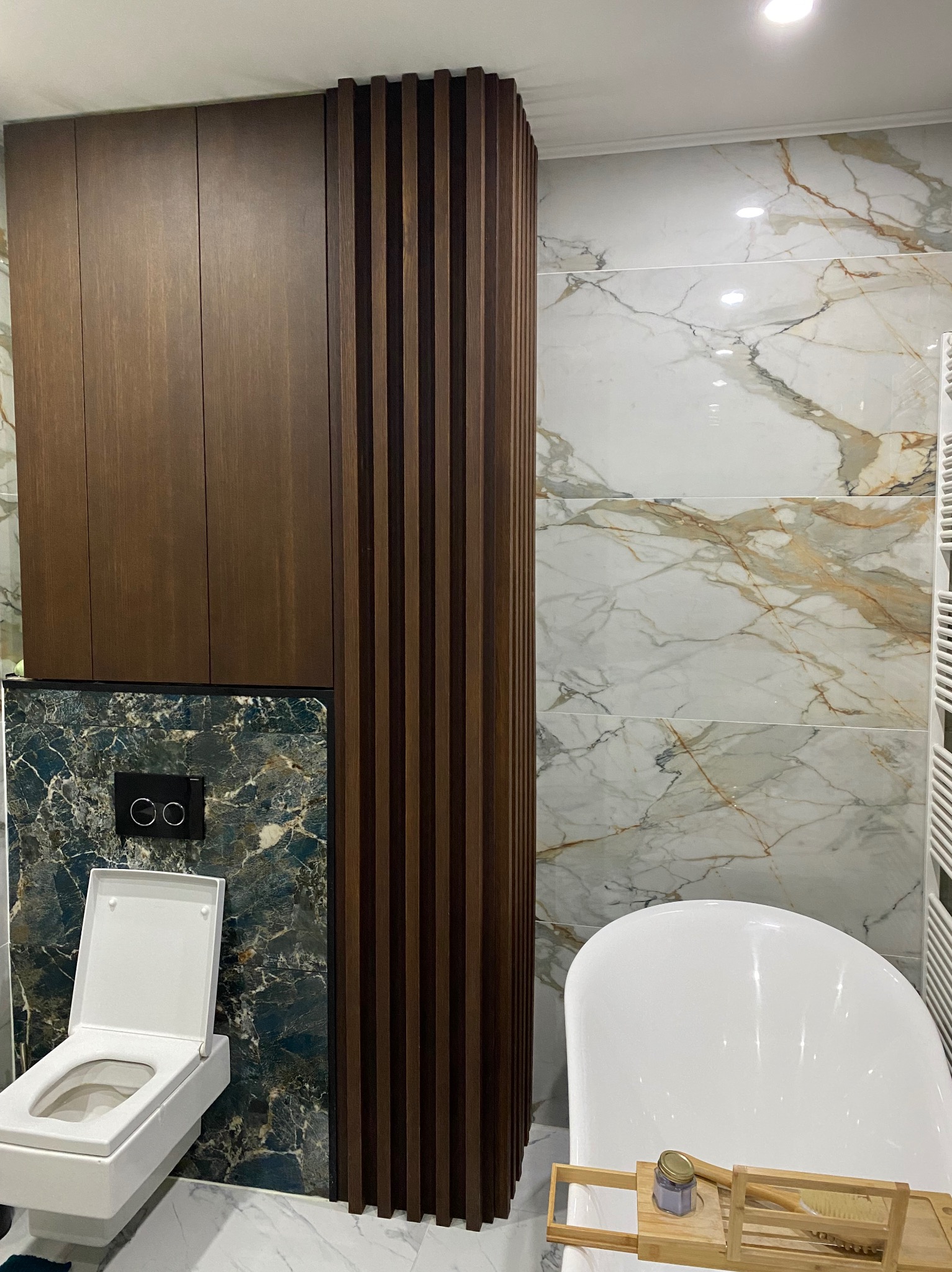 Nowoczesna łazienka z marmurowymi płytkami, zabudową z ciemnego drewna, wiszącą toaletą i białą wanną z drewnianą półką.