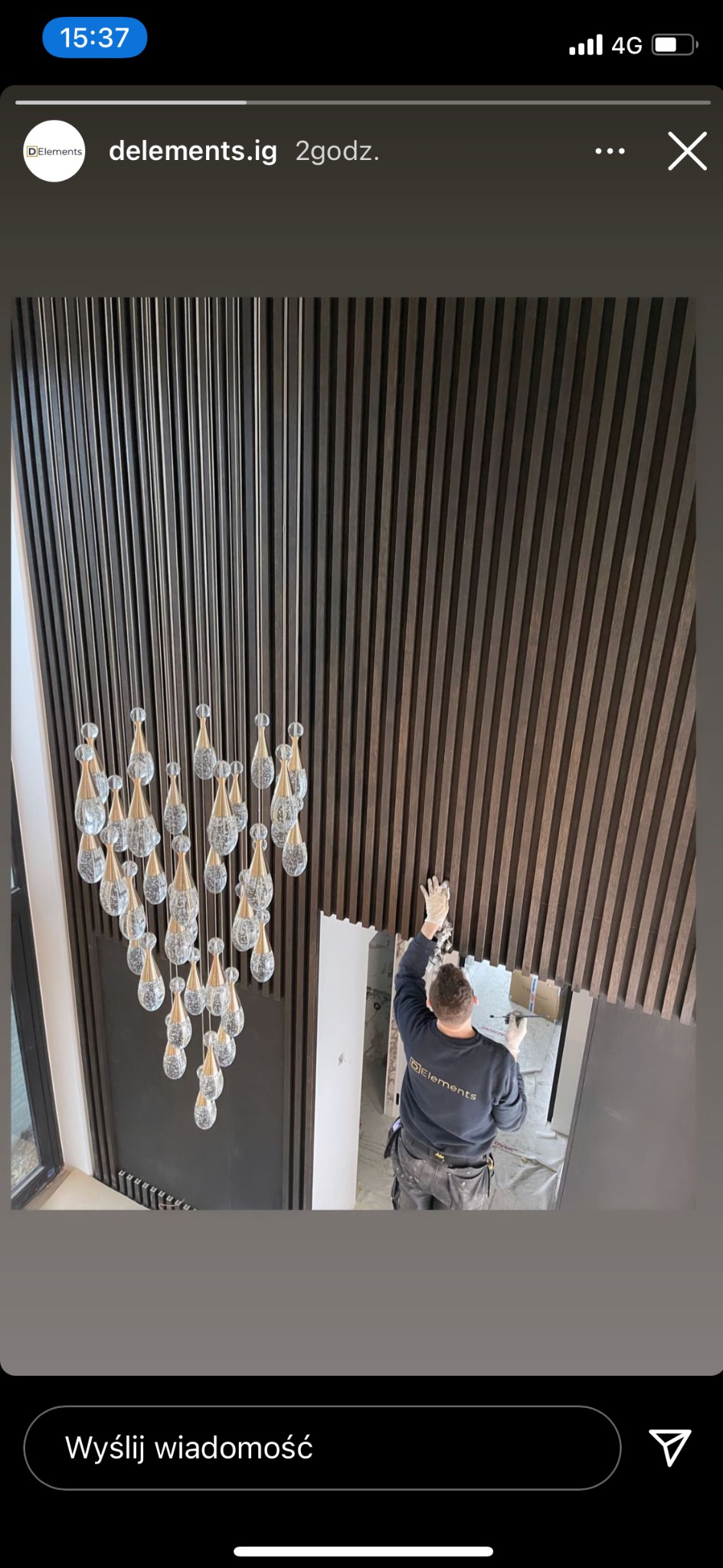 Montaż paneli ściennych z ciemnego drewna w wysokim pomieszczeniu, widoczny pracownik w rękawicach na tle jasnych ścian i wiszący żyrandol z kryształkami