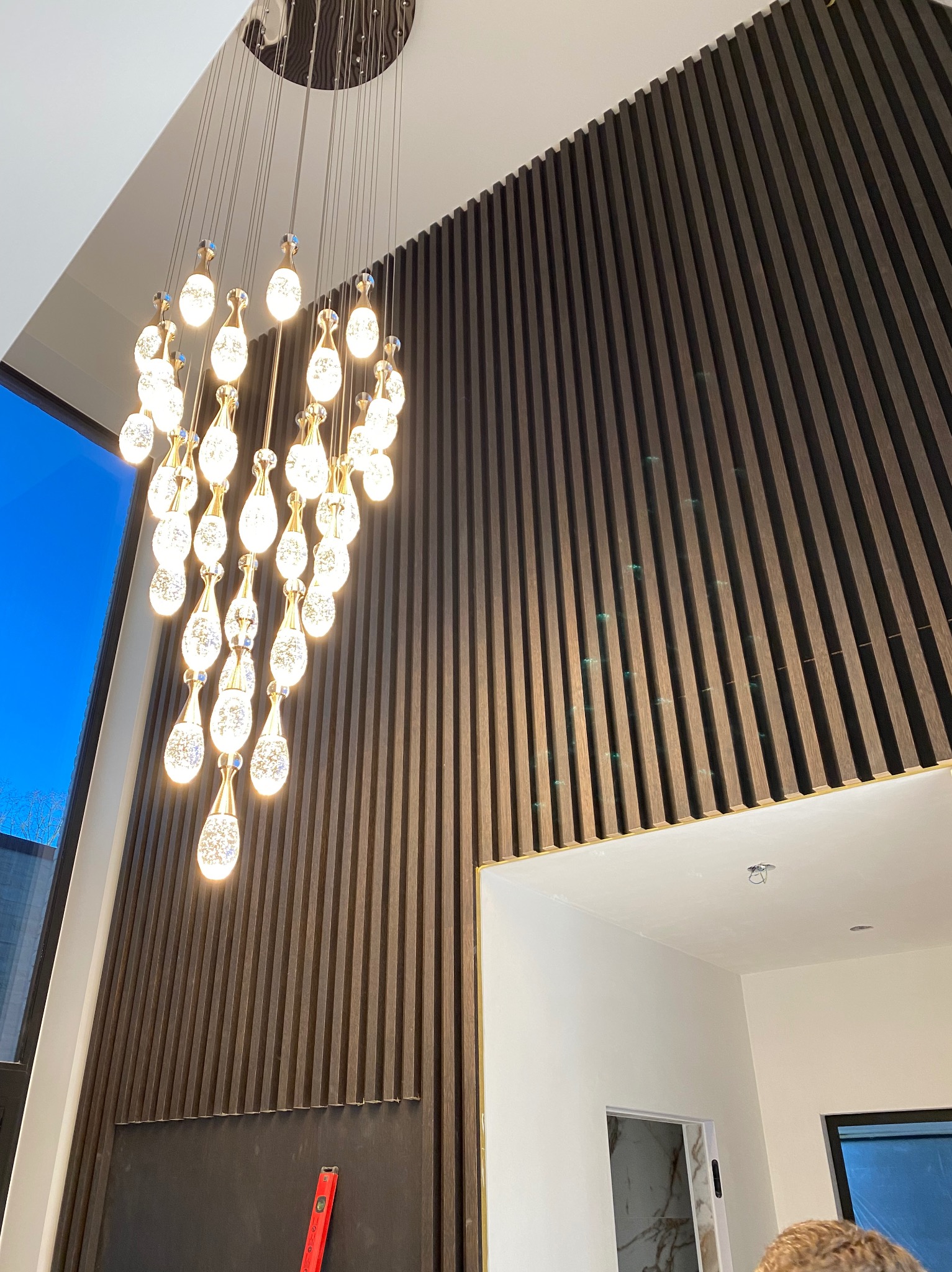 Nowoczesny żyrandol z kryształowymi elementami w kształcie łez, zawieszony na tle ściany z paneli drewnianych, widoczny fragment sufitu i okna z niebieskim niebem w tle.