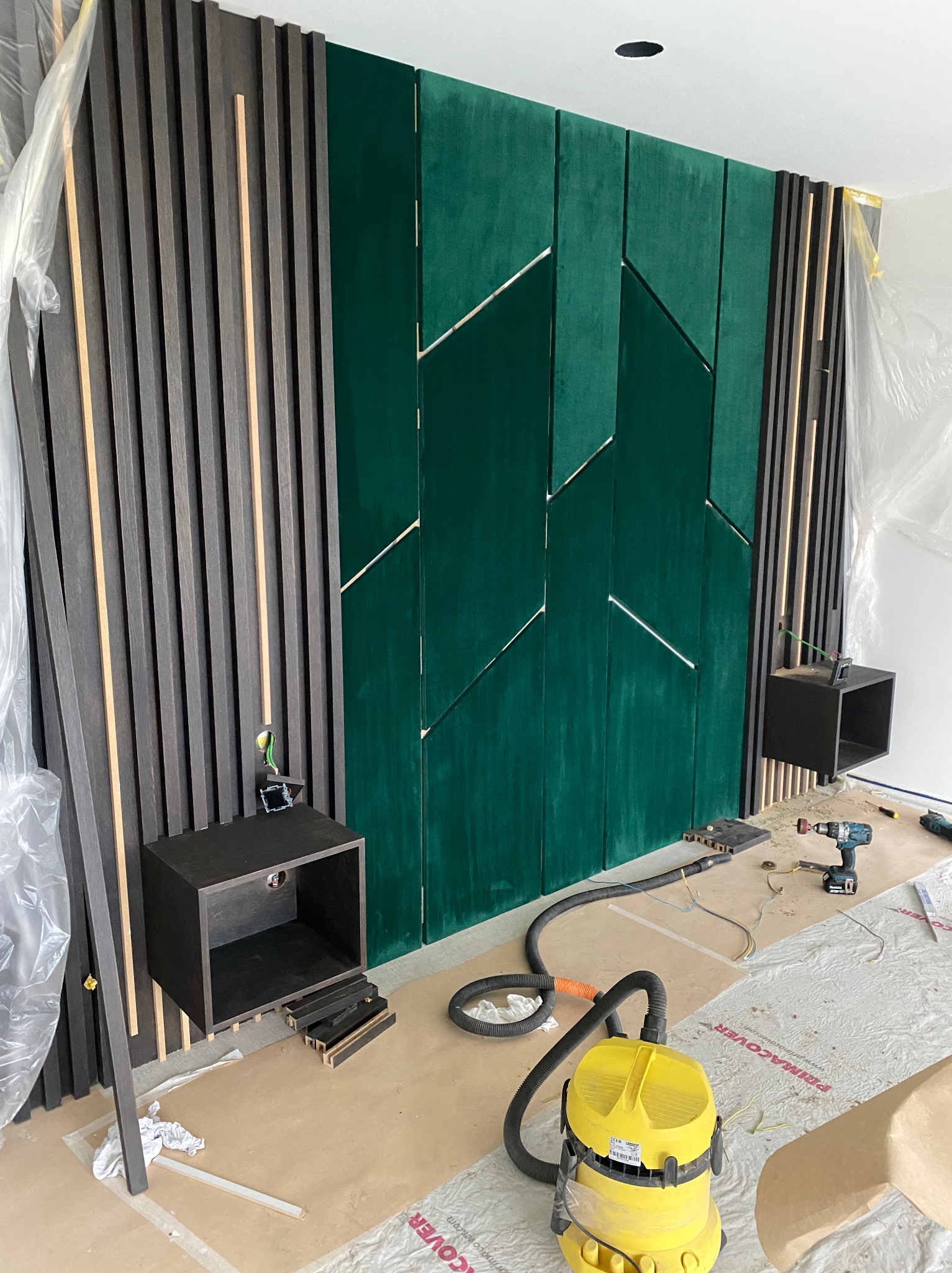 Montaż panelu ściennego z ciemnego drewna i zielonego weluru z geometrycznym wzorem, widoczne narzędzia i szafki nocne w trakcie instalacji, zabezpieczona podłoga.