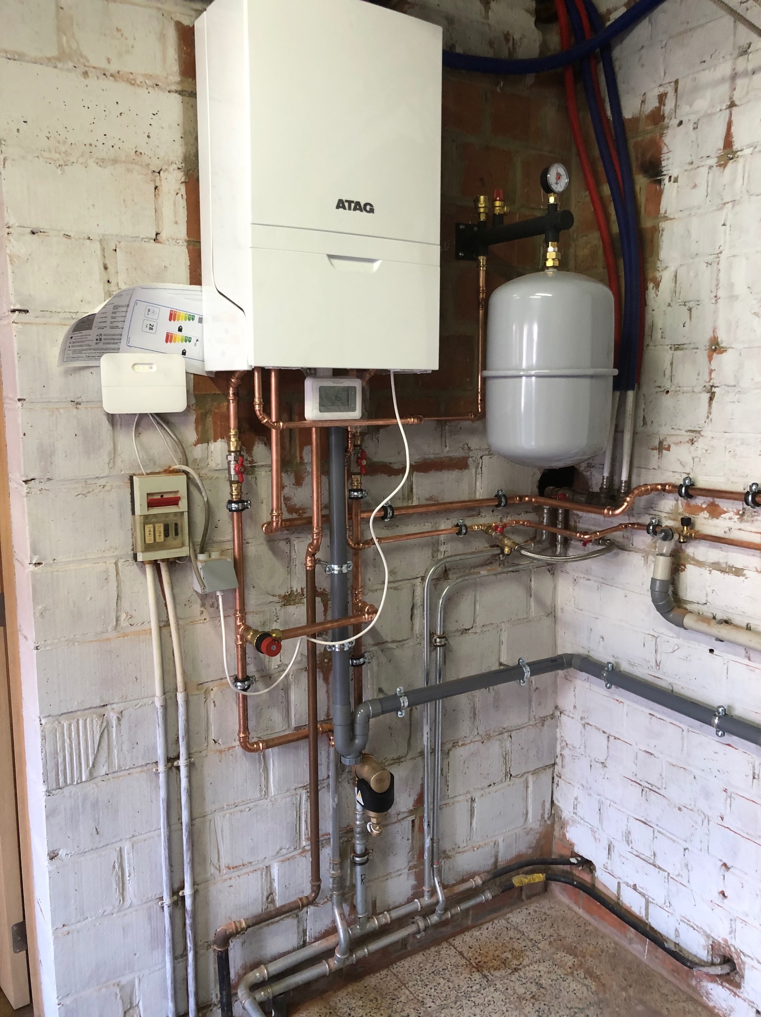 Nowoczesny, biały piec gazowy marki ATAG zamontowany na białej, ceglanej ścianie. Widoczne miedziane rury, szary zbiornik wyrównawczy, manometr i instalacja elektryczna.