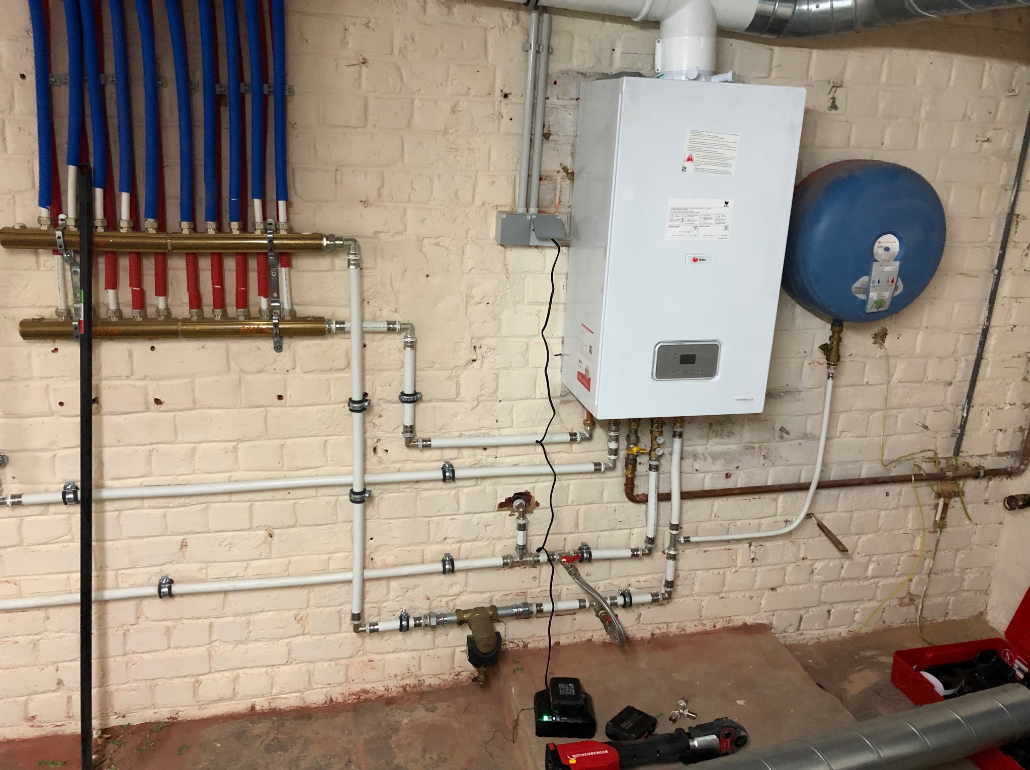 Instalacja centralnego ogrzewania z rozdzielaczem, piecem gazowym i naczyniem przeponowym na tle malowanej ceglanej ściany. Na podłodze widoczne narzędzia instalacyjne.