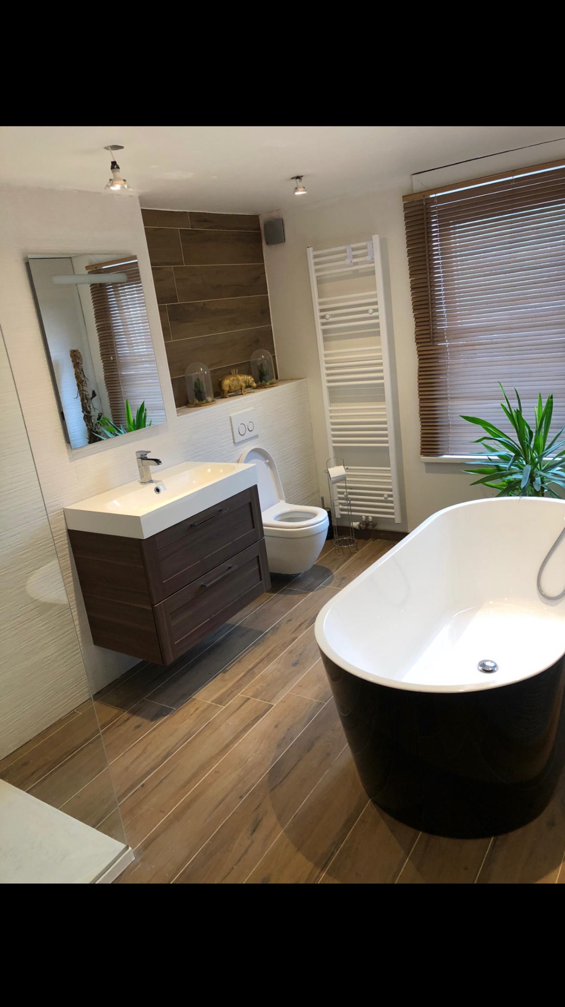 Nowoczesna łazienka z drewnopodobnymi płytkami, czarną wanną wolnostojącą, szafką pod umywalką z ciemnego drewna i grzejnikiem drabinkowym.