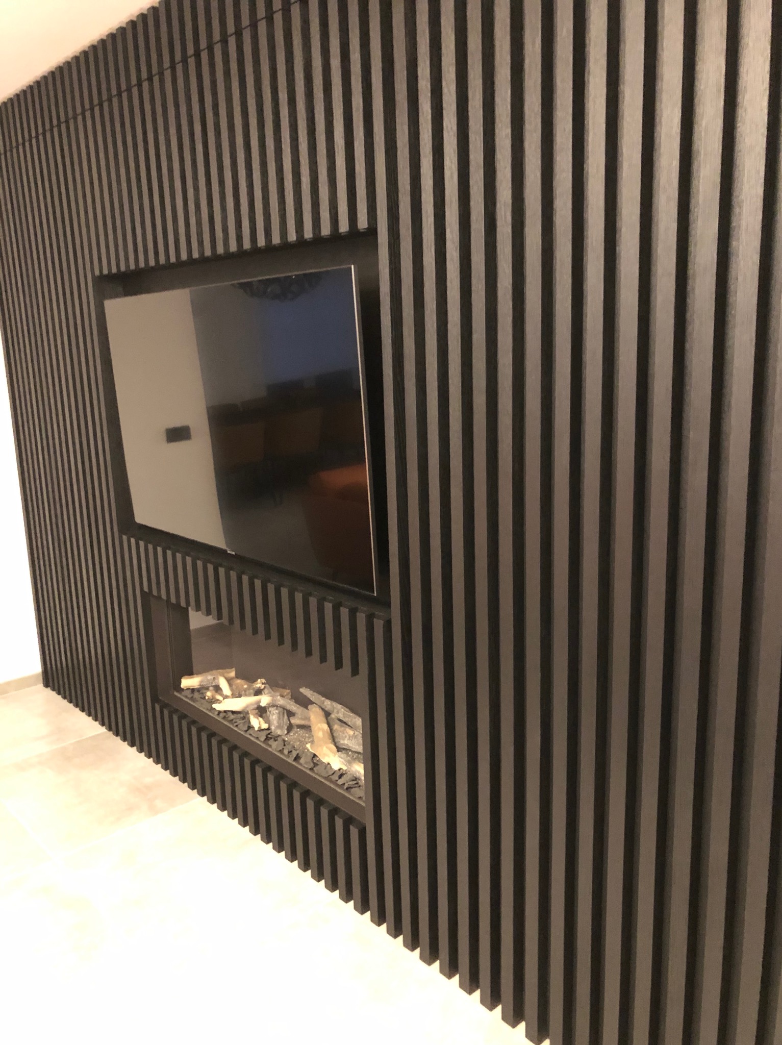 Ściana z czarnych, pionowych lameli z wnęką na telewizor i imitacją kominka z dekoracyjnymi polanami.