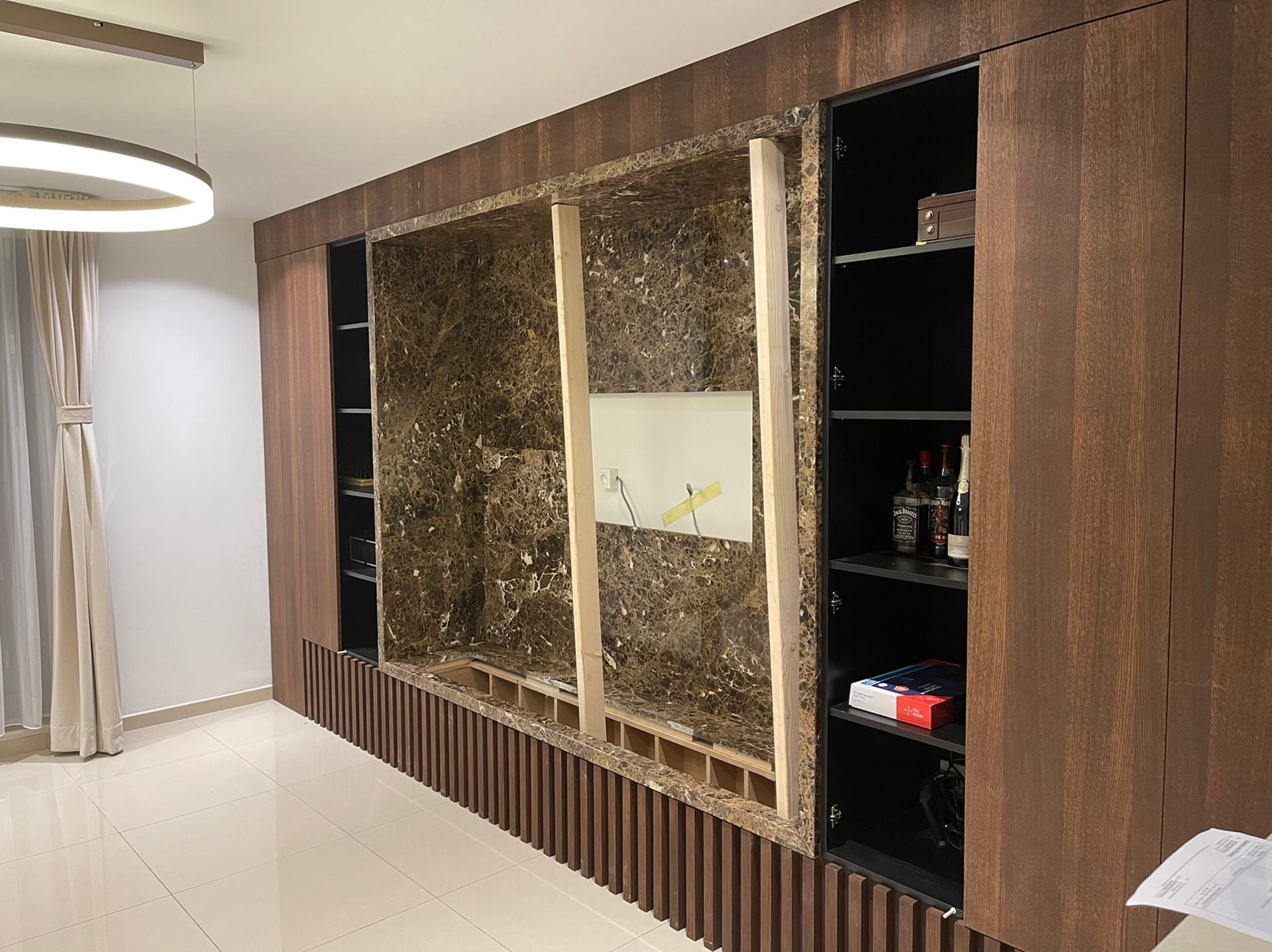 Wnęka w zabudowie meblowej z imitacją marmuru w trakcie przygotowania do montażu, widoczne drewniane podpory i fragment instalacji elektrycznej, na półkach obok butelki alkoholu.