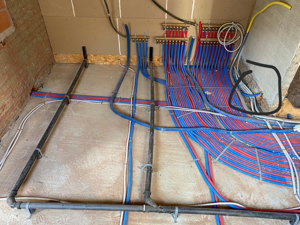 Instalacja ogrzewania podłogowego w trakcie realizacji: liczne czerwono-niebieskie rury prowadzone do rozdzielaczy na ścianie, czarne rury kanalizacyjne przy podłodze, surowa ściana z cegły i płyty...