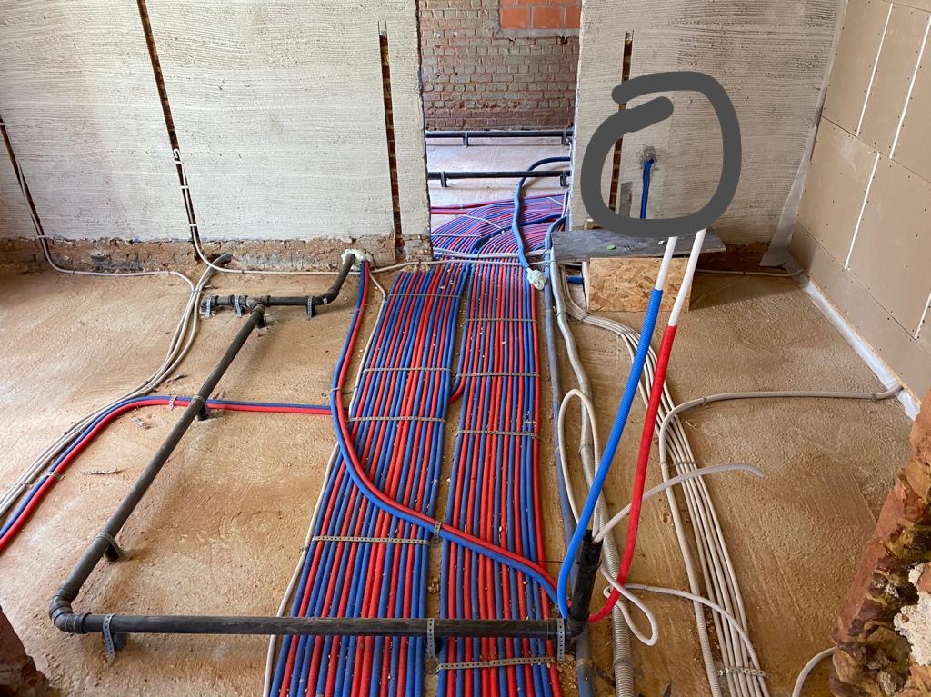 Instalacja ogrzewania podłogowego w trakcie realizacji, widoczne rury grzewcze w kolorach czerwonym i niebieskim, ściany w stanie surowym, widoczne instalacje elektryczne i wodne.