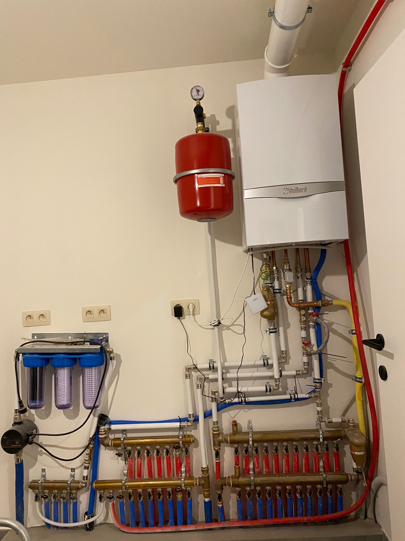 Kompletna instalacja centralnego ogrzewania z piecem gazowym Vaillant, czerwonym zbiornikiem wyrównawczym, rozdzielaczami mosiężnymi i filtrami wody, zamontowana na ścianie w kotłowni.