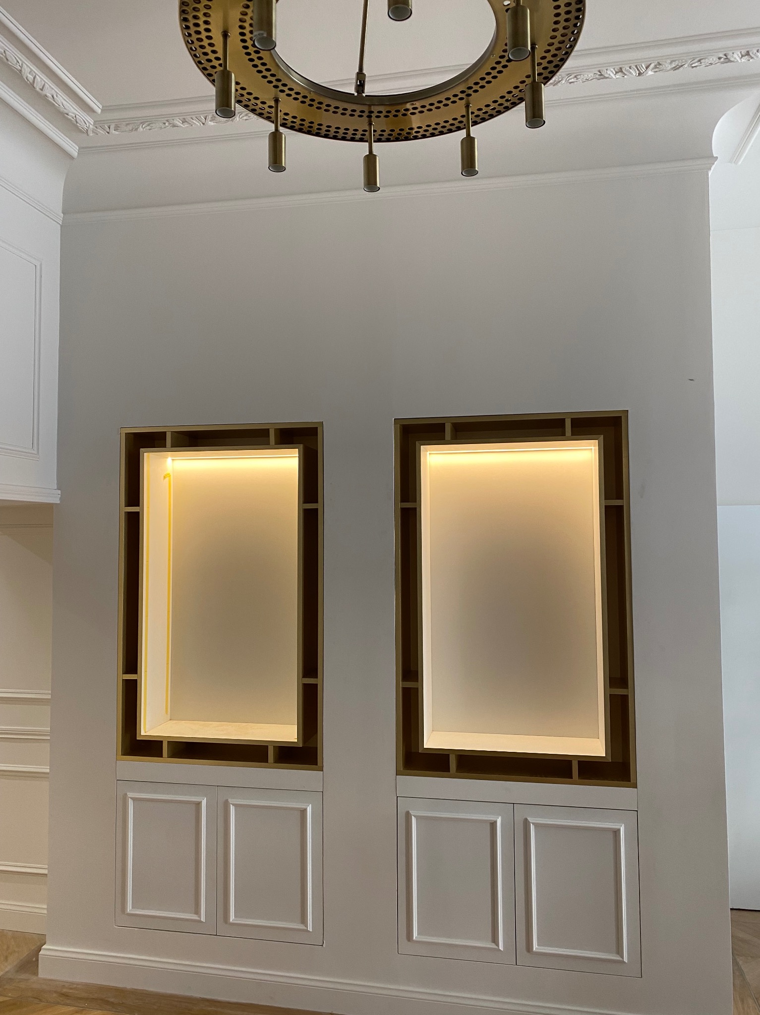 Dwa podświetlane wnęki z półkami i szafkami w dolnej części, umieszczone w białej ścianie z dekoracyjną listwą sufitową i złotym żyrandolem.