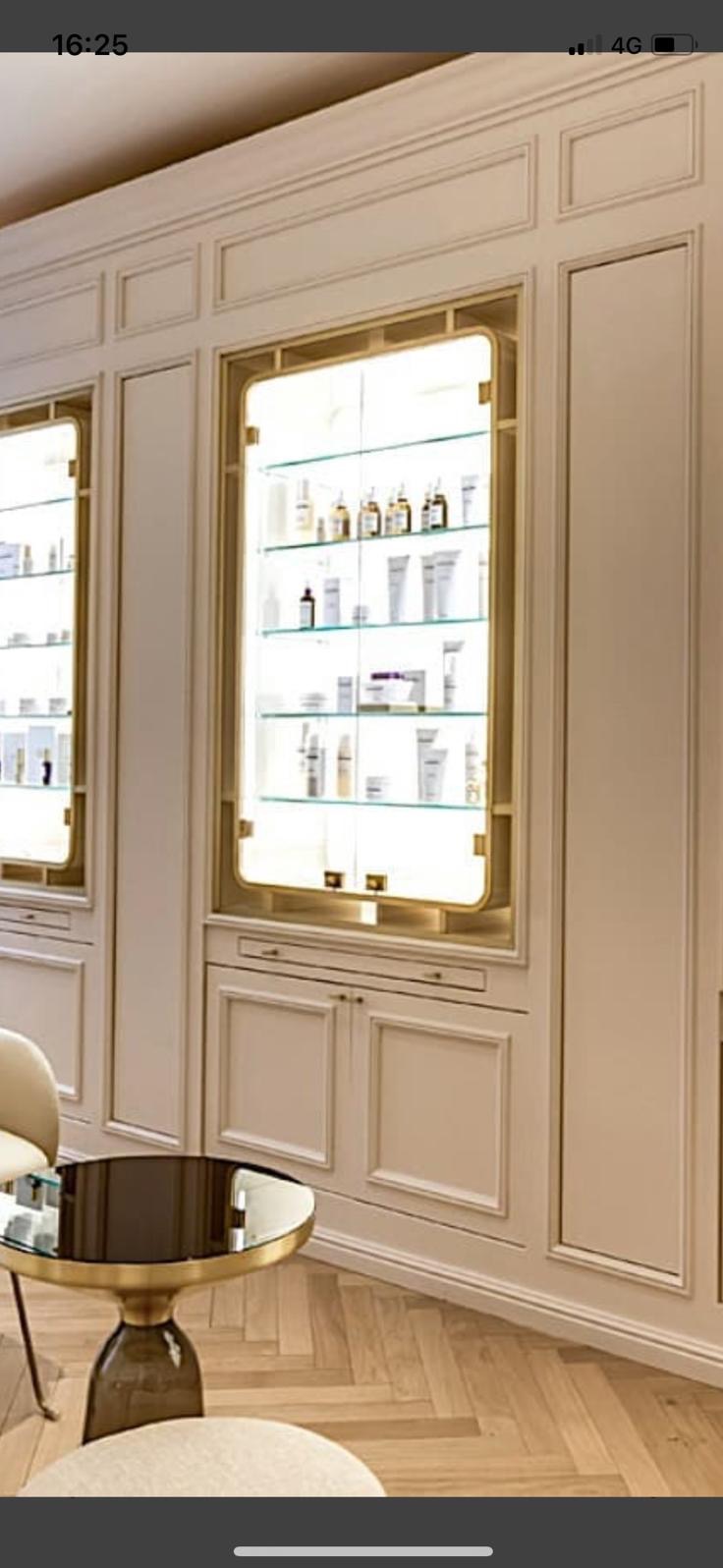 Ściana z wbudowanymi, podświetlanymi witrynami z kosmetykami i kremami w eleganckim salonie kosmetycznym, widoczny fragment szklanego stolika i parkietu.