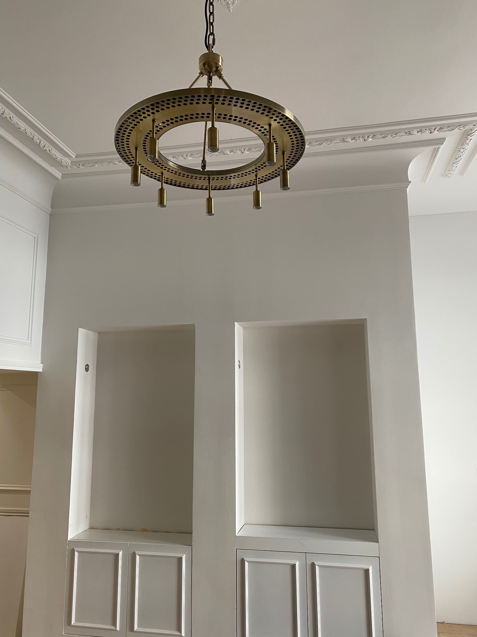 Wnętrze z białymi ścianami, sztukaterią sufitową i wnękami z szafkami, oświetlone przez okrągły, złoty żyrandol z punktowymi otworami.