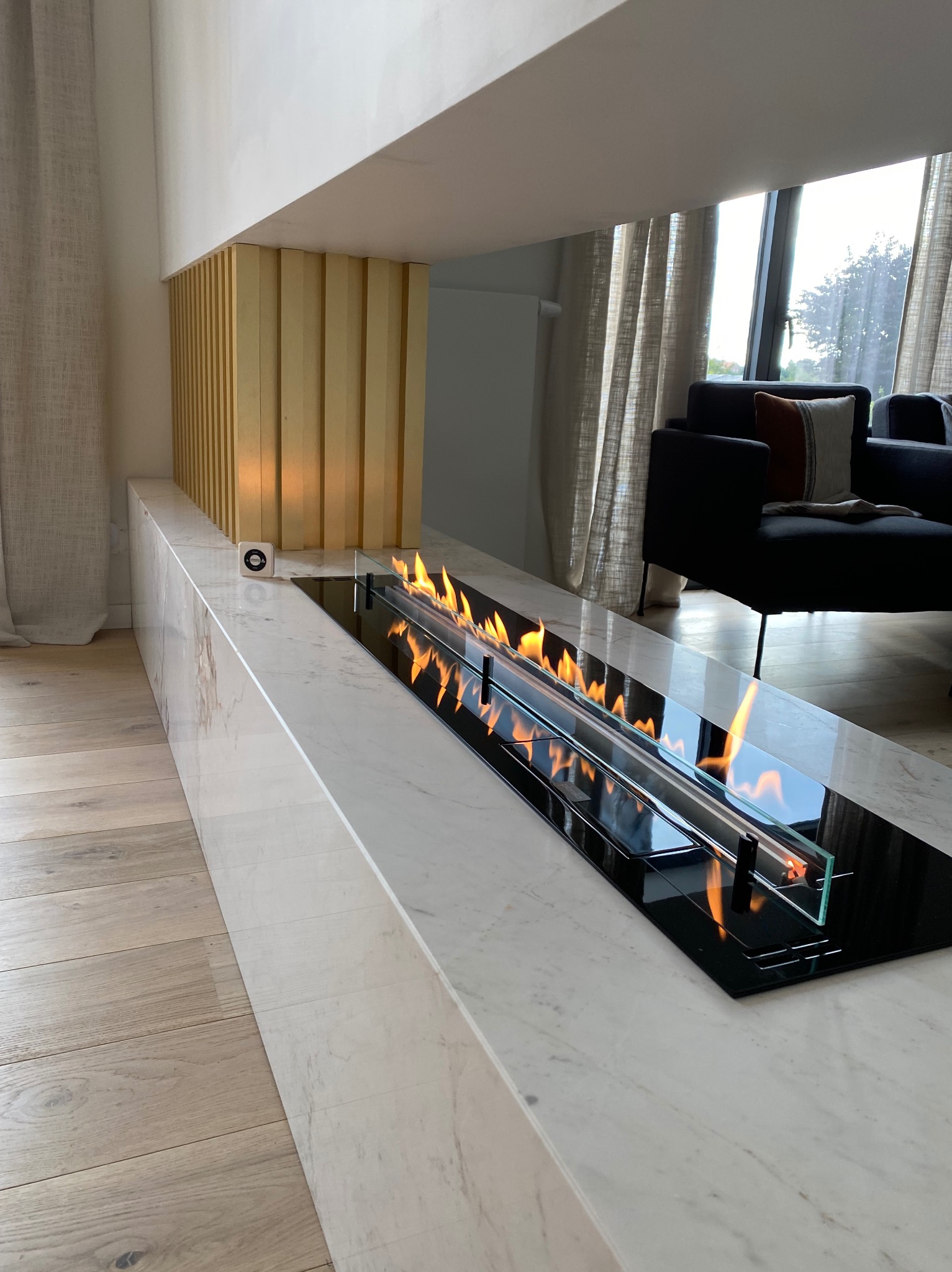 Nowoczesny biokominek z paleniskiem w obudowie z białego marmuru, z widocznym sterownikiem, na tle drewnianej podłogi i zasłon w salonie.