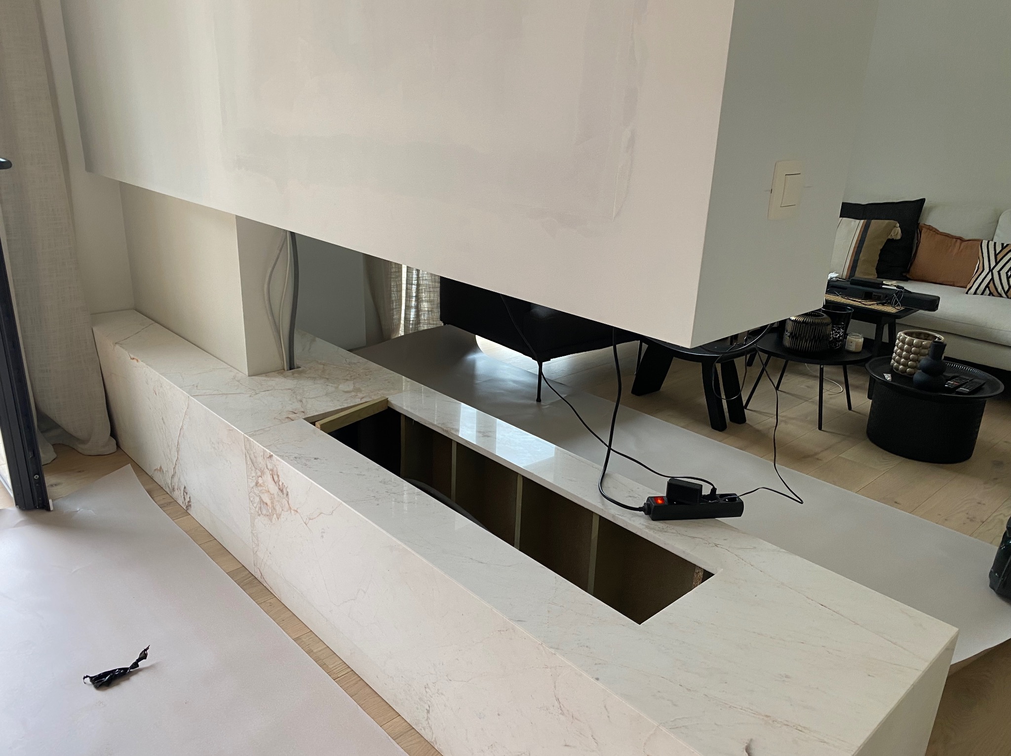 Nowoczesny kominek z marmurowym wykończeniem w trakcie montażu, zabezpieczony folią malarską, widoczne kable i listwa zasilająca, w tle salon.