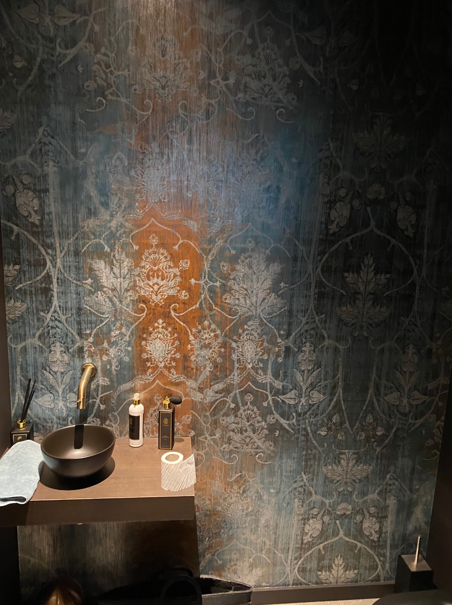 Elegancka umywalka nablatowa w ciemnej kolorystyce, złota bateria i dekoracyjna tapeta w odcieniach błękitu i brązu z motywem roślinnym, uzupełniona o stylowe akcesoria łazienkowe.