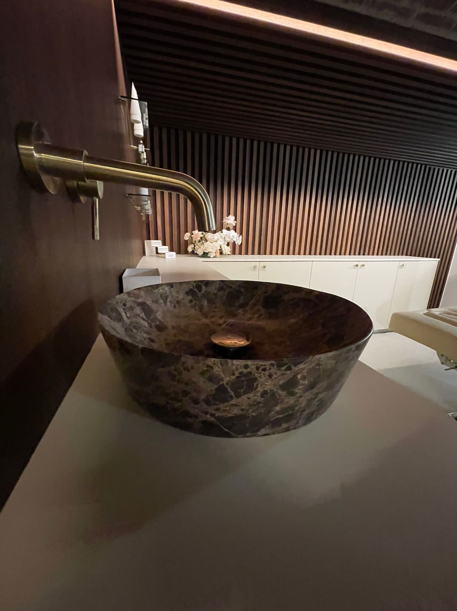 Marmurkowa umywalka nablatowa z mosiężną baterią na tle drewnianej ściany i białych szafek w nowoczesnej łazience.