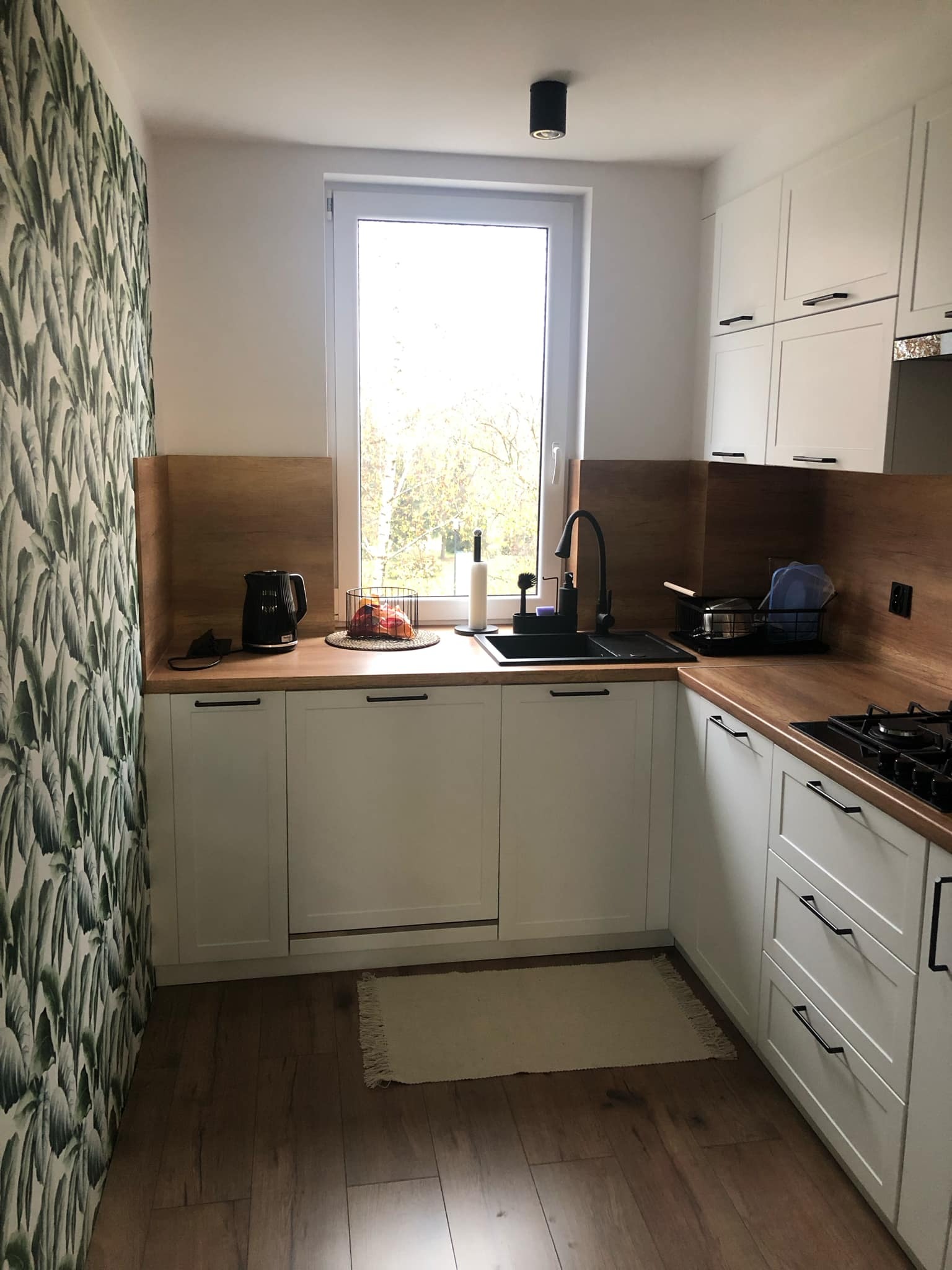 Jasna kuchnia z białymi szafkami, drewnianym blatem i tapetą w liście bananowca, czarny zlew i bateria, widok na zewnątrz przez okno.