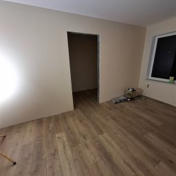 Art-Fliz - Pokój w trakcie prac wykończeniowych: panele podłogowe ułożone, ściany pomalowane na beżowo, widoczne narzędzia przy oknie, surowy otwór drzwiowy bez ościeżnicy.