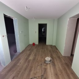 Art-Fliz - Wnętrze w trakcie prac wykończeniowych: zielone ściany, panele podłogowe, przygotowane otwory drzwiowe, widoczny kabel zwinięty na szpuli na podłodze.