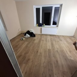 Art-Fliz - Pokój po remoncie z nową podłogą laminowaną, widoczne profile do montażu płyt karton-gips oraz osoba robiąca zdjęcie telefonem.