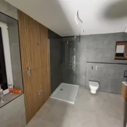 Nowoczesna łazienka w odcieniach szarości z drewnianymi panelami ściennymi, kabiną prysznicową typu walk-in i podwieszaną toaletą, widoczne narzędzia i materiały budowlane na parapecie okna.