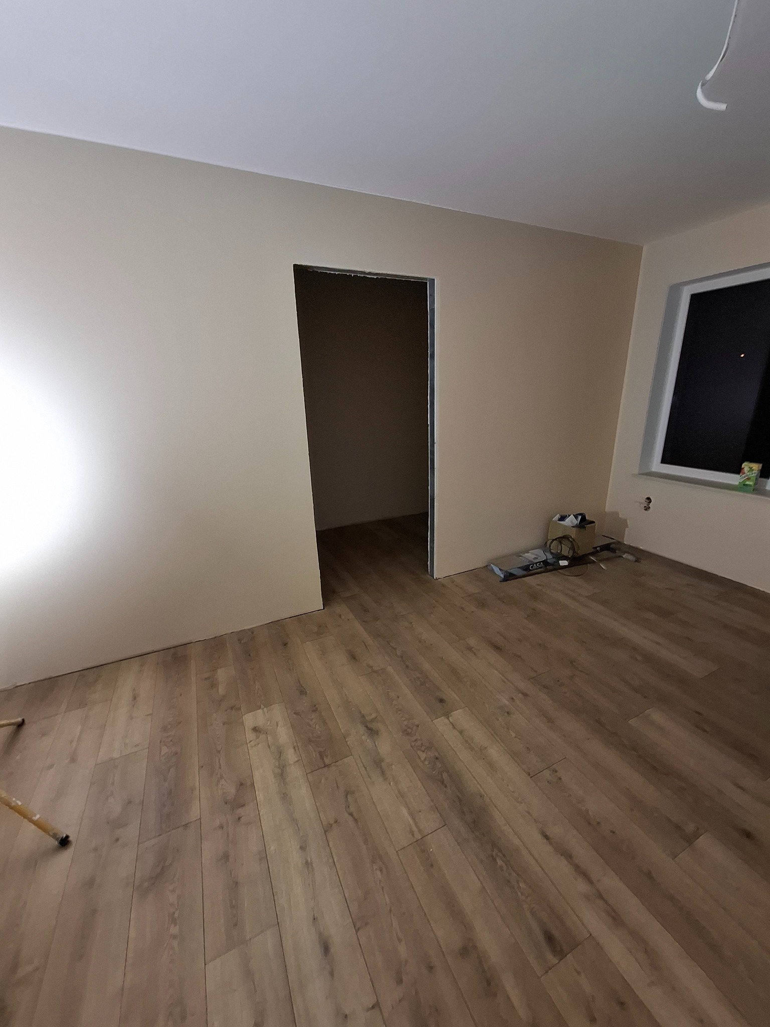 Pokój w trakcie prac wykończeniowych: panele podłogowe ułożone, ściany pomalowane na beżowo, widoczne narzędzia przy oknie, surowy otwór drzwiowy bez ościeżnicy.