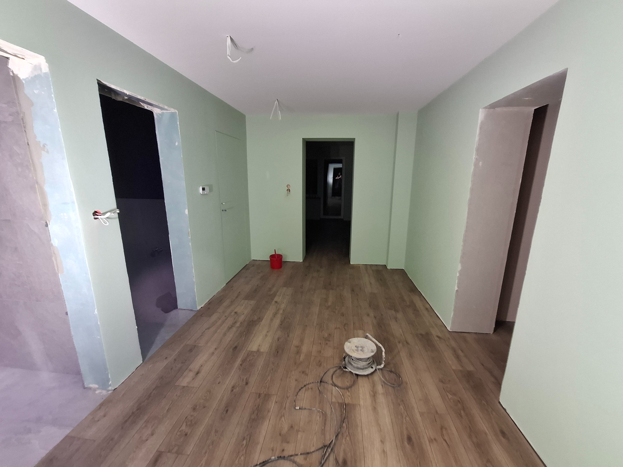 Wnętrze w trakcie prac wykończeniowych: zielone ściany, panele podłogowe, przygotowane otwory drzwiowe, widoczny kabel zwinięty na szpuli na podłodze.