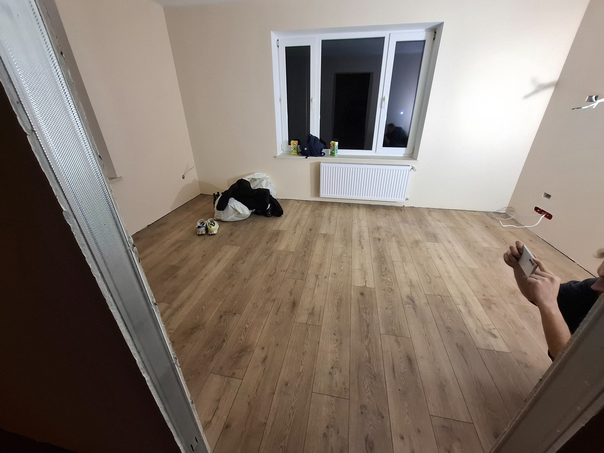 Pokój po remoncie z nową podłogą laminowaną, widoczne profile do montażu płyt karton-gips oraz osoba robiąca zdjęcie telefonem.