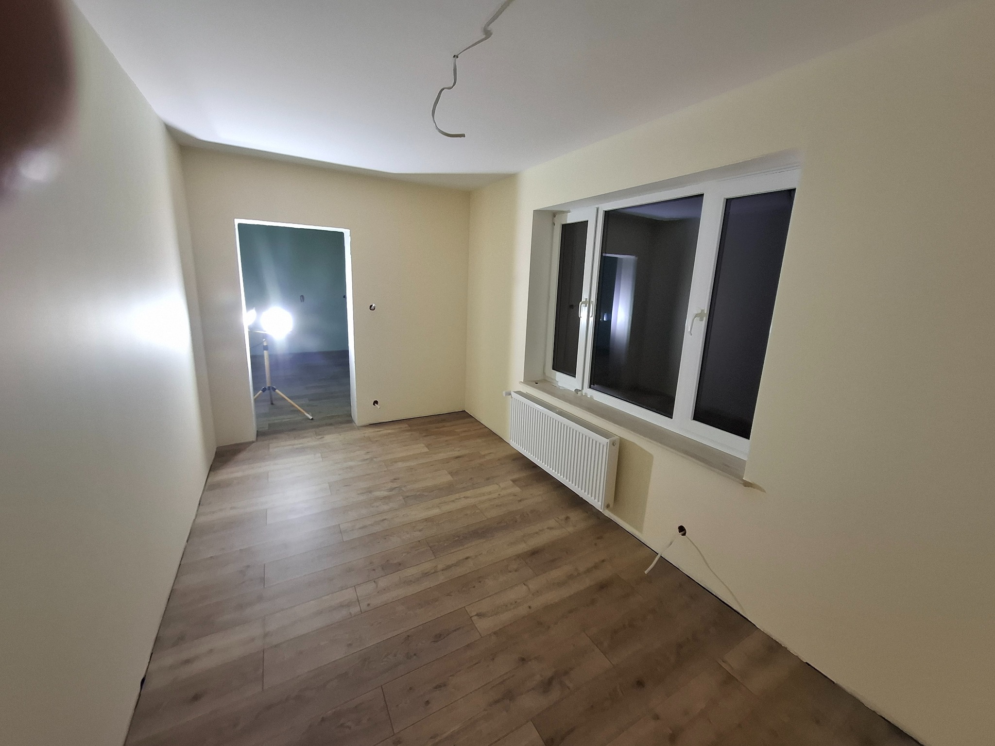 Wnętrze pokoju po malowaniu ścian na jasny kolor, z widocznymi panelami podłogowymi, grzejnikiem pod oknem i wystającymi kablami elektrycznymi ze ściany oraz wiszącym kablem z sufitu.