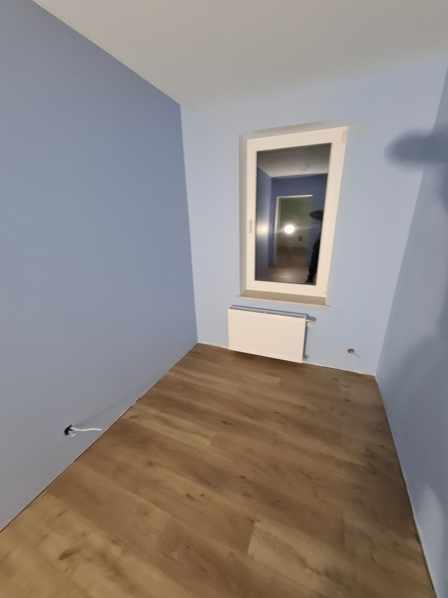 Pomieszczenie po malowaniu na niebiesko z panelami podłogowymi, widoczny grzejnik pod oknem i wystające kable ze ściany.