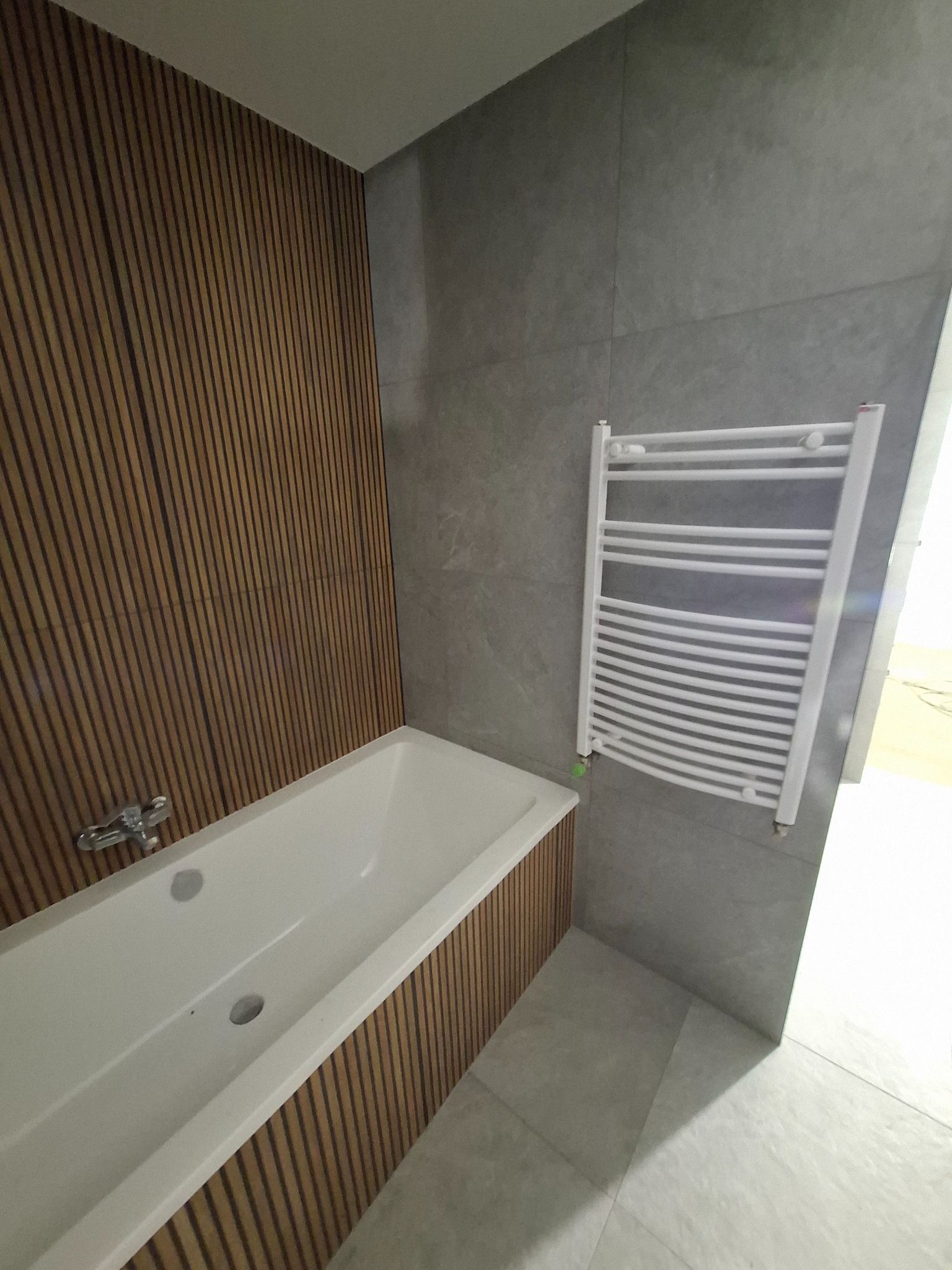 Wanna obudowana drewnianymi panelami pionowymi, szara płytka na ścianie z grzejnikiem drabinkowym.