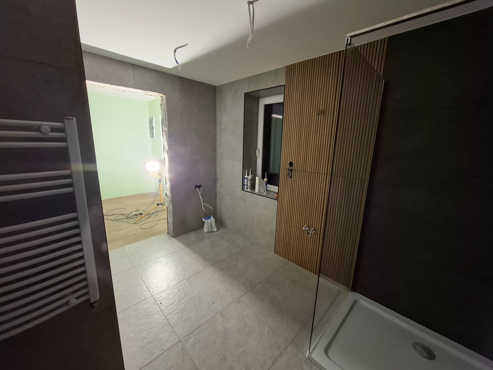 Łazienka w trakcie remontu: szare płytki, drewniana zabudowa, kabina prysznicowa z przezroczystą szybą, widoczne przewody elektryczne i narzędzia, surowy stan ścian w przejściu do innego...