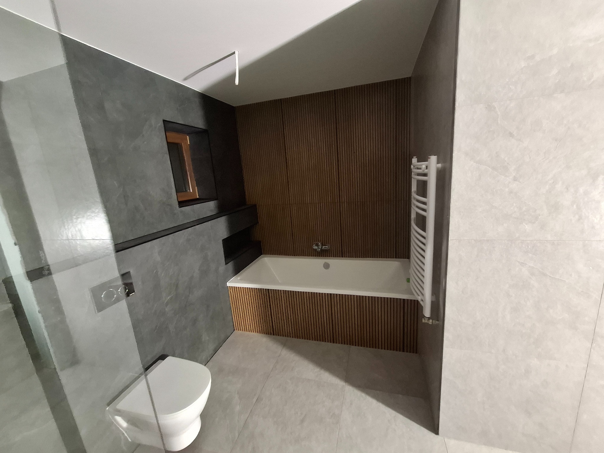 Nowoczesna łazienka z szarą ścianą imitującą beton, drewnianą zabudową wanny, wiszącą toaletą i grzejnikiem drabinkowym