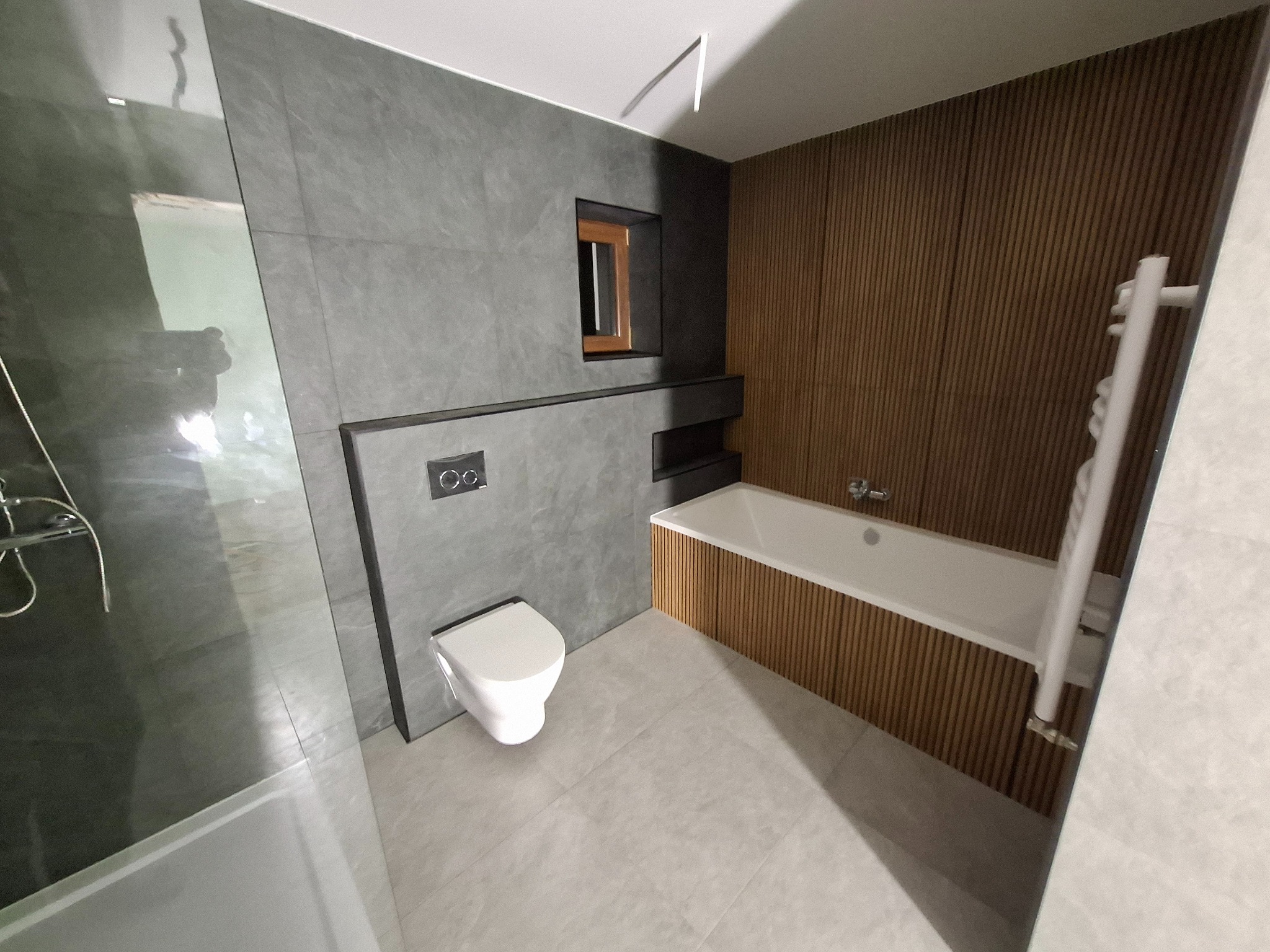 Nowoczesna łazienka z szarymi płytkami imitującymi beton, drewnianymi panelami przy wannie i minimalistyczną toaletą wiszącą. Małe okienko z drewnianą ramą dodaje charakteru.