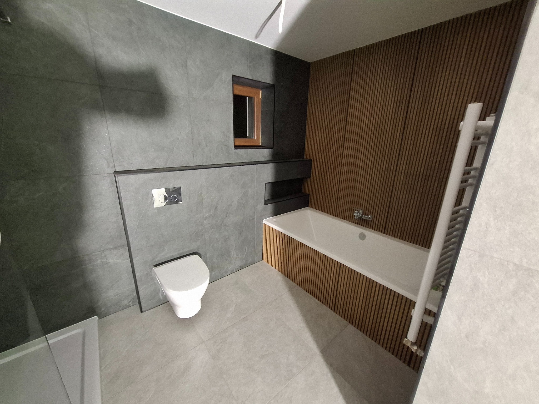 Nowoczesna łazienka z szarymi płytkami imitującymi kamień, drewnianymi panelami przy wannie, podtynkową toaletą i małym oknem w drewnianej ramie.