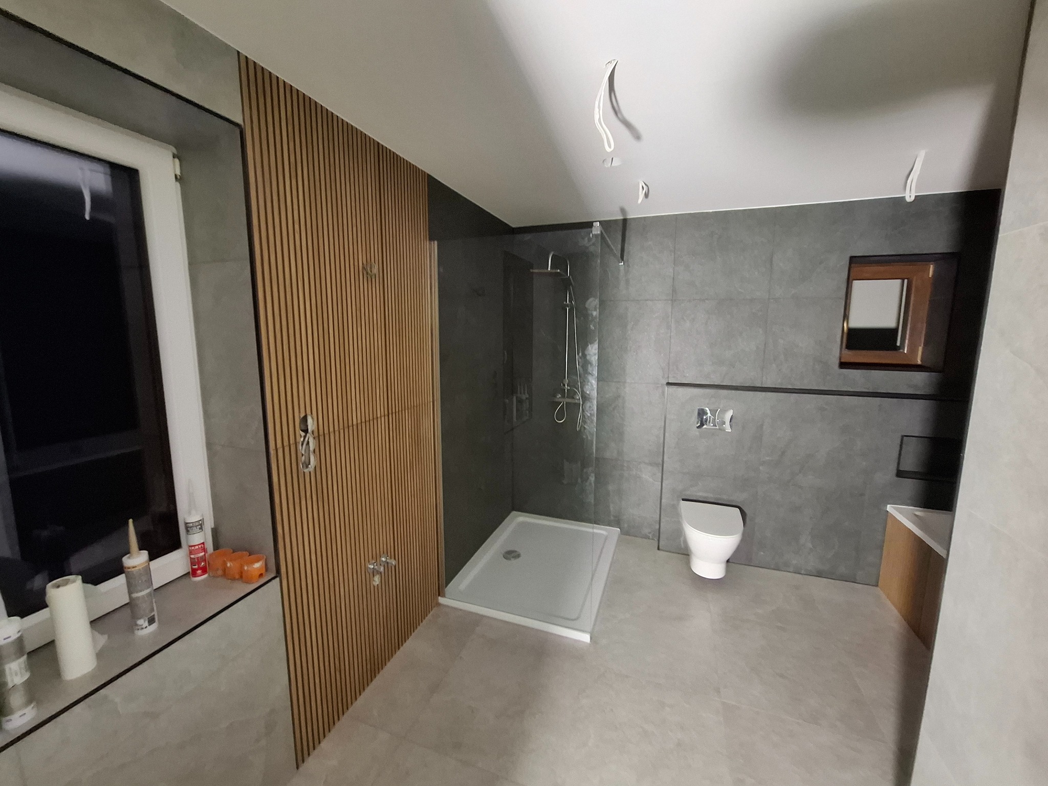 Nowoczesna łazienka w odcieniach szarości z drewnianymi panelami ściennymi, kabiną prysznicową typu walk-in i podwieszaną toaletą, widoczne narzędzia i materiały budowlane na parapecie okna.