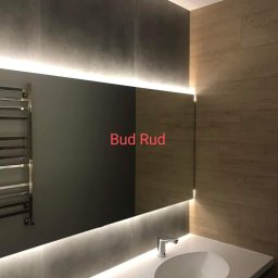 Bud Rud - Nowoczesna łazienka z podświetlanym lustrem, szare płytki na ścianie i drewnopodobne panele, chromowany grzejnik drabinkowy po lewej stronie, biała umywalka z dwiema bateriami.