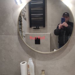 Bud Rud - Okrągłe lustro w łazience odbija grzejnik drabinkowy, białą umywalkę z złotą baterią, dozownik mydła i rolkę papieru toaletowego. W lustrze widoczny mężczyzna robiący zdjęcie telefonem.