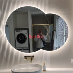 Bud Rud - Nowoczesna łazienka z owalną umywalką nablatową, złotą baterią i podświetlanym lustrem; w odbiciu lustra widoczna pralka i suszarka w zabudowie oraz osoba robiąca zdjęcie.
