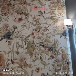 Bud Rud - Ściana wyłożona płytkami z motywem kwiatów i papug w łazience, oświetlona lampą, widoczny fragment lustra.