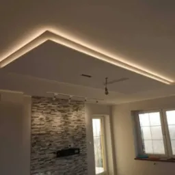 Nowoczesny salon z podwieszanym sufitem oświetlonym taśmą LED, dekoracyjną ścianą z kamienia i oknami z widokiem na zewnątrz.