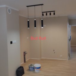 Bud Rud - Pomalowane na beżowo wnętrze z drewnopodobną podłogą, widoczne wiszące lampy, puszki po farbie i czarny worek przy ścianie.