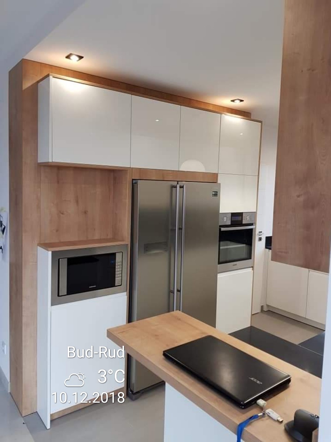 Nowoczesna kuchnia z białymi, lakierowanymi szafkami górnymi i zabudową z forniru drewnopodobnego, obejmującą mikrofalówkę i lodówkę side-by-side; na blacie leży czarny laptop Acer.