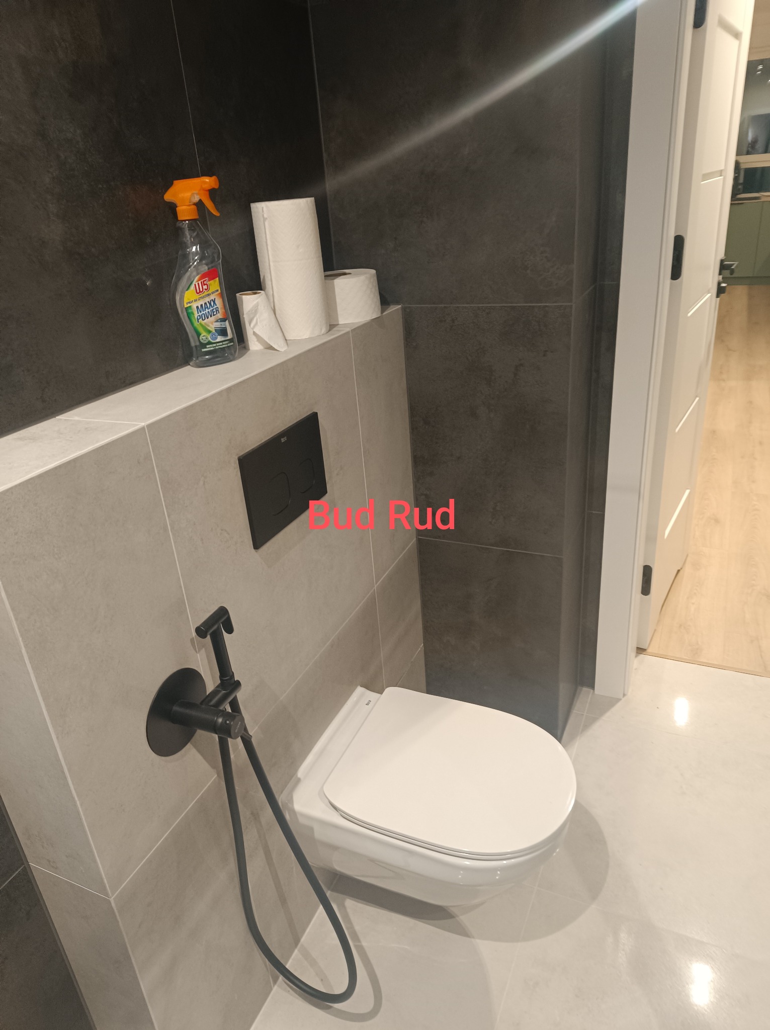 Nowoczesna toaleta wisząca z czarną armaturą bidetową na tle szarych płytek imitujących beton, uzupełniona o dozownik mydła i rolki papieru toaletowego.