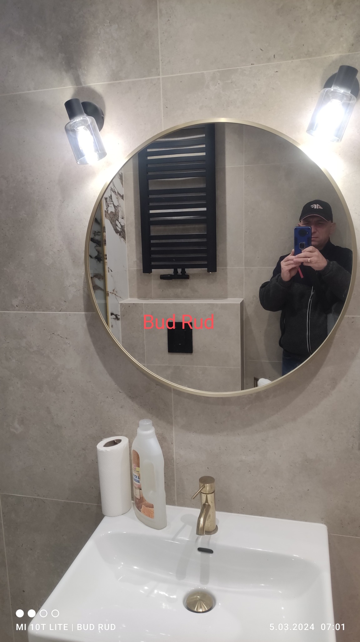 Okrągłe lustro w łazience odbija grzejnik drabinkowy, białą umywalkę z złotą baterią, dozownik mydła i rolkę papieru toaletowego. W lustrze widoczny mężczyzna robiący zdjęcie telefonem.